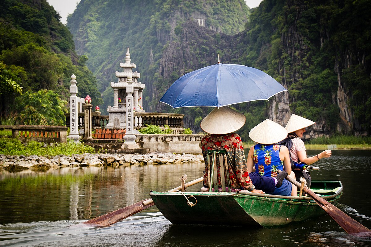 Women in traditional conical hats rowing a boat past a riverside temple in Tam Coc with karst cliffs